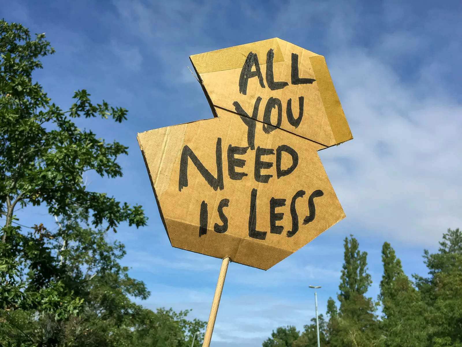 Cartel de cartón con el mensaje «All you need is less» frente a un cielo azul y árboles